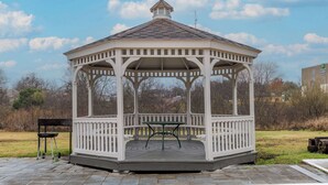 Un gazebo dalla forma esagonale con tetto a punta, circondato da un prato e alberi.