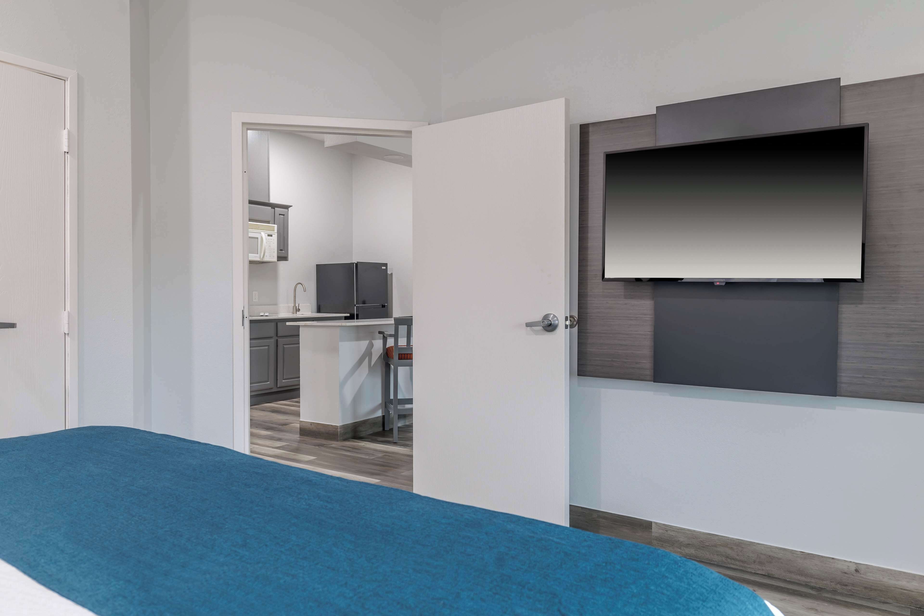 A modern hotel room with a bed, a TV mounted on the wall, and a view into the kitchen area.