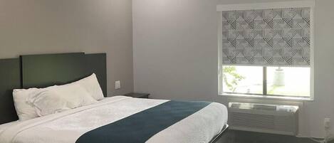 A modern hotel room with a large bed, a bedside table, a window with patterned blinds, and a wall-mounted air conditioning unit.