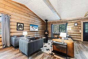 Interior de una cabaña de madera con sofá, mesa, chimenea y ventana con cortinas.