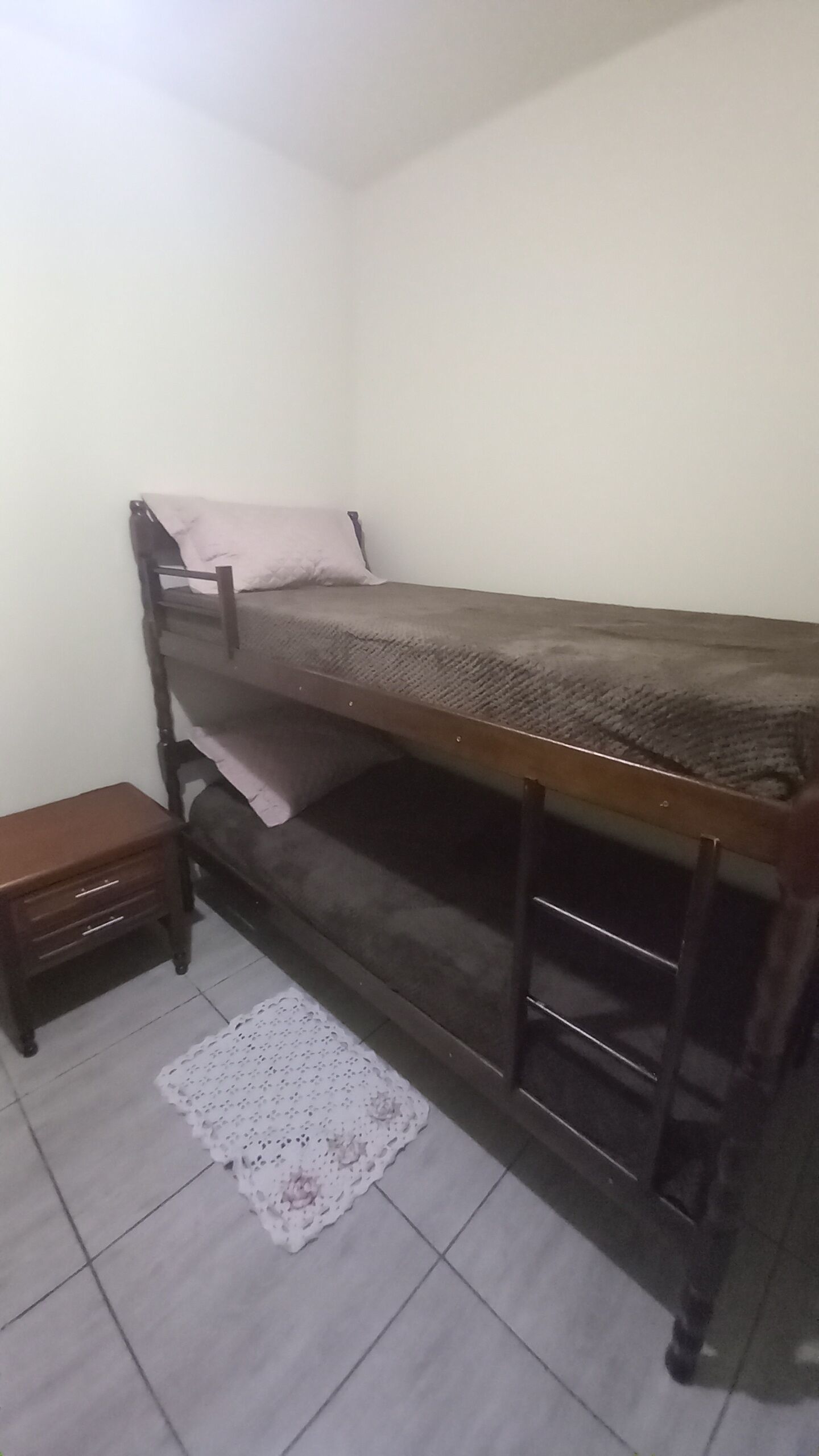 Una cama individual con estructura de madera y un colchón. Hay una mesita de noche de madera con cajones y un mantel de encaje en el suelo.