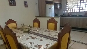 Ein Essbereich mit einem für sechs Personen gedeckten Tisch, umgeben von Holzstühlen mit gelben Kissen. Im Hintergrund ist eine Küche mit Holdelementen und einem Kühlschrank zu sehen.