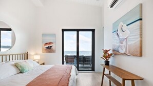 A bedroom with a bed, a bench, a table, and a view of the sea through a sliding door. - Beachfront Luxury Suite #19 at THE BEACH HOUSE (Campbell River)