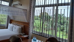 Une chambre d’hôtel avec un lit, une table de chevet et une vue sur la verdure à travers des fenêtres grillagées.