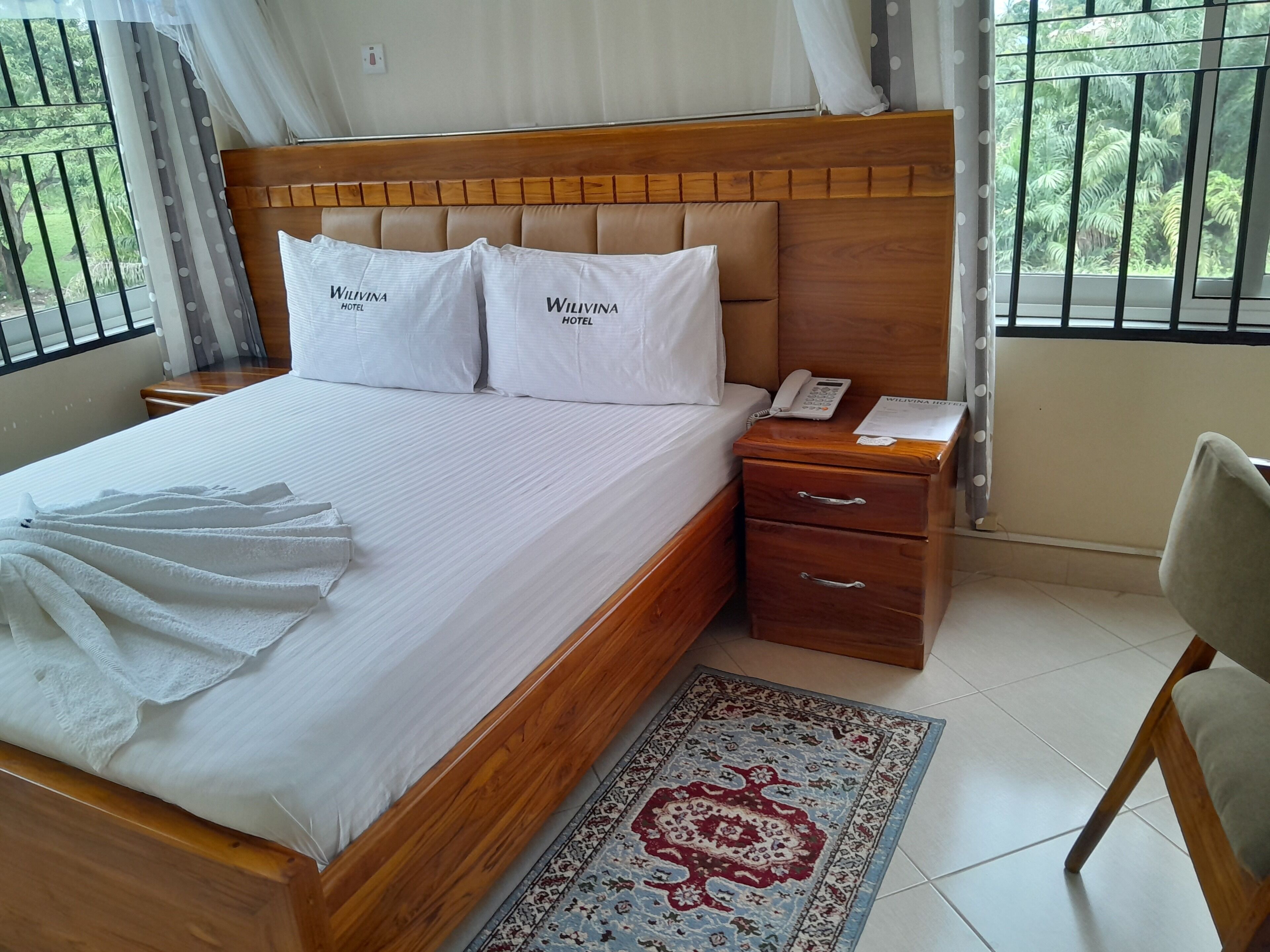 A neatly made bed with white bedding and pillows, a wooden headboard, a bedside table with a phone and a menu, and a patterned rug on a tiled floor.