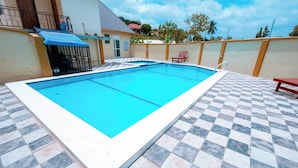 Une piscine rectangulaire dont le fond est décoré d’un motif à carreaux, entourée d’un petit muret et bordée d’une zone abritée par une marquise bleue.