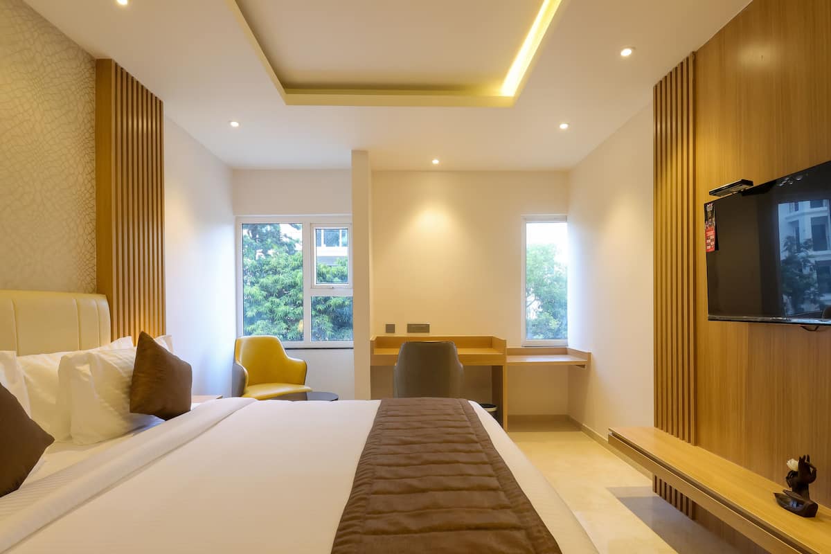 Habitación de hotel con una cama grande, un escritorio, una silla, un televisor y ventanas con vista a vegetación.