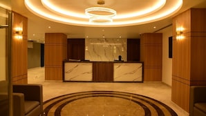 A hotel reception area with a marble reception desk, wooden paneling, a circular carpet, and a chandelier. - Hotel Grand Serene Mysore (Mysore)