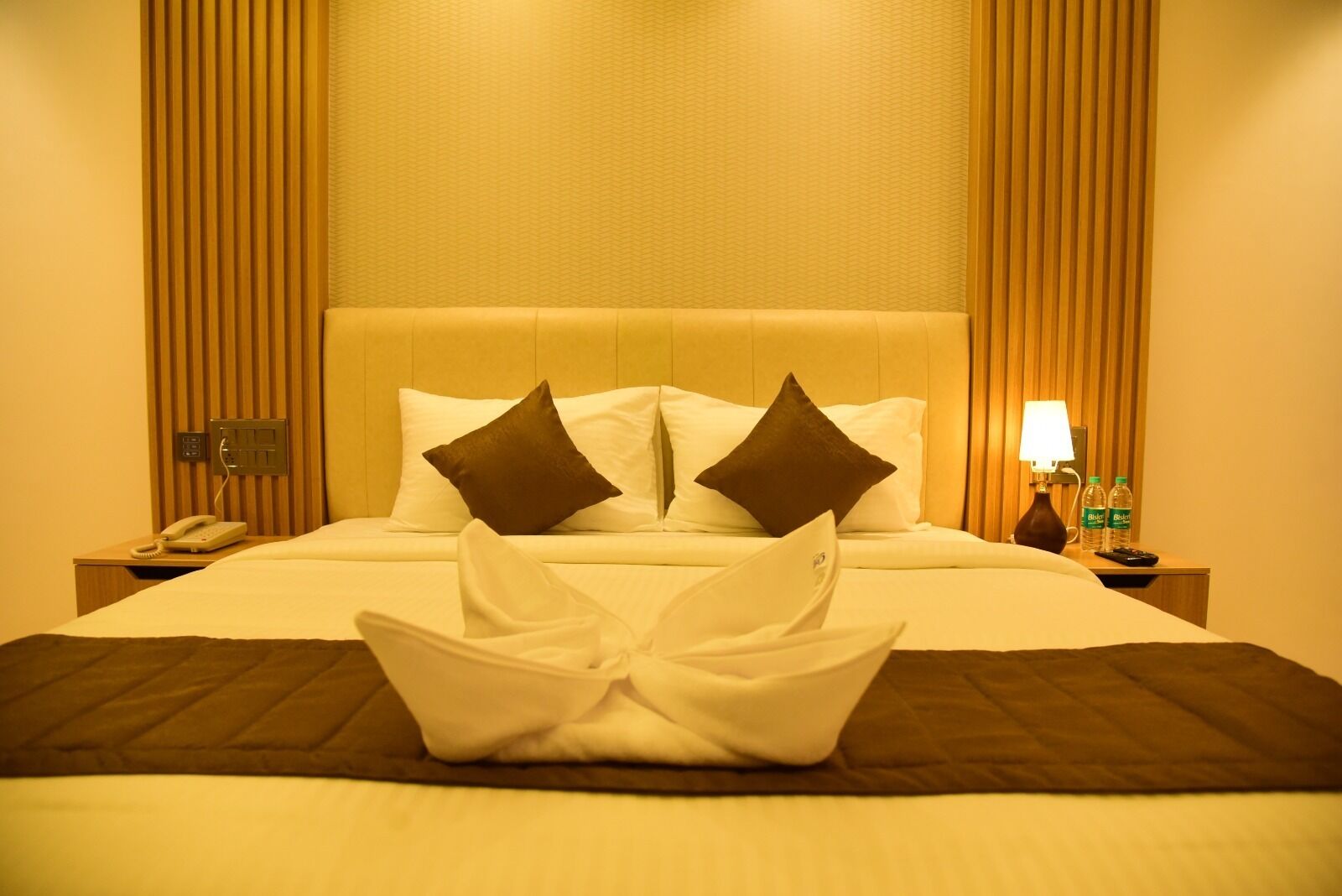 Une chambre d’hôtel avec un lit, des oreillers, une table de chevet avec une lampe, un téléphone et deux bouteilles d’eau.