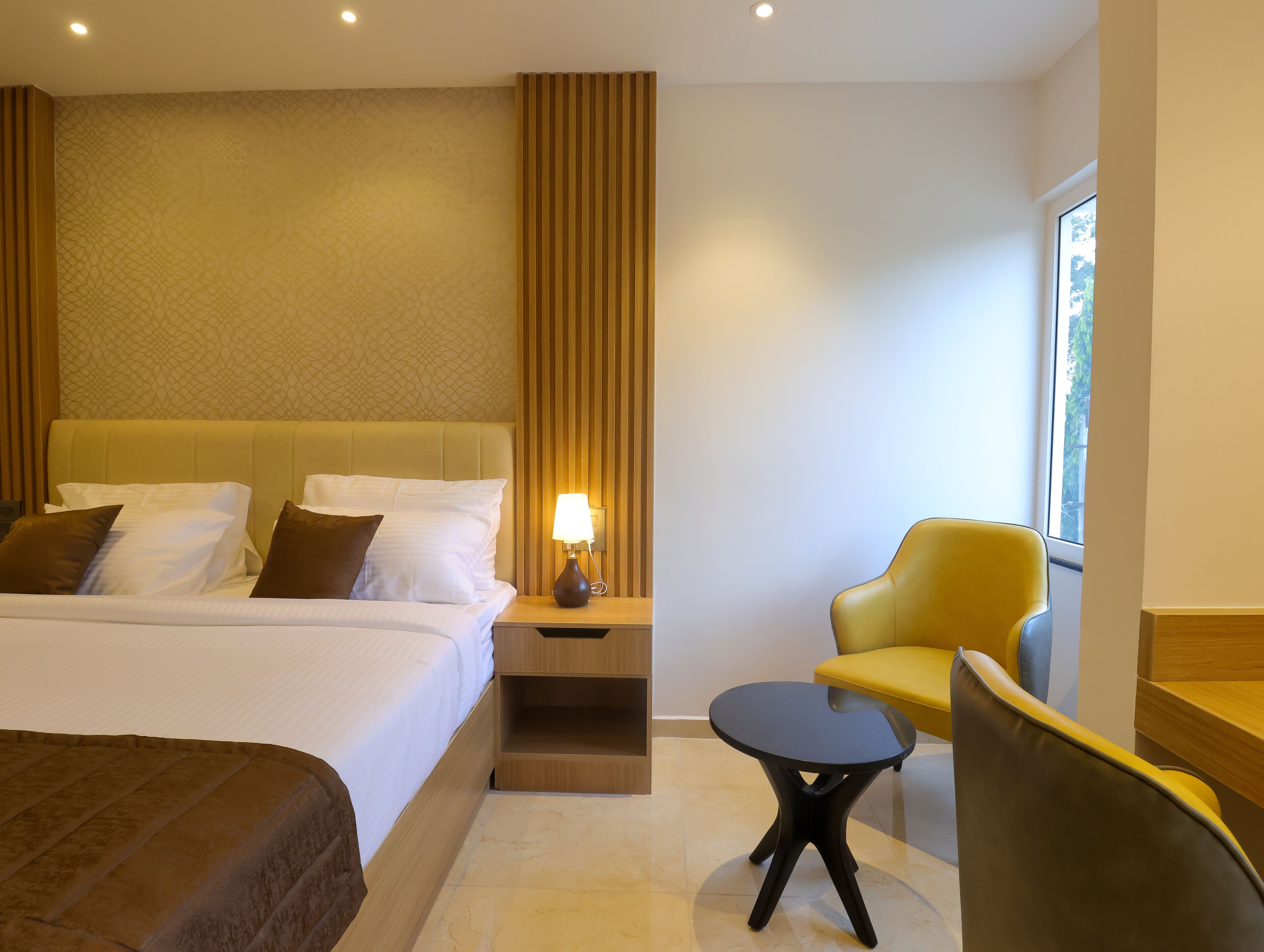 A hotel room with a large bed, a bedside table with a lamp, a small round table, and a yellow armchair.