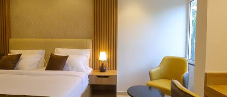 A hotel room with a large bed, a bedside table with a lamp, a small round table, and a yellow armchair.