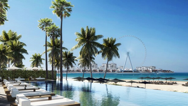 Un espace aménagé au bord de la piscine, avec des chaises longues et des parasols, des palmiers, un ciel bleu azur et, au loin, une grande roue et des bâtiments.