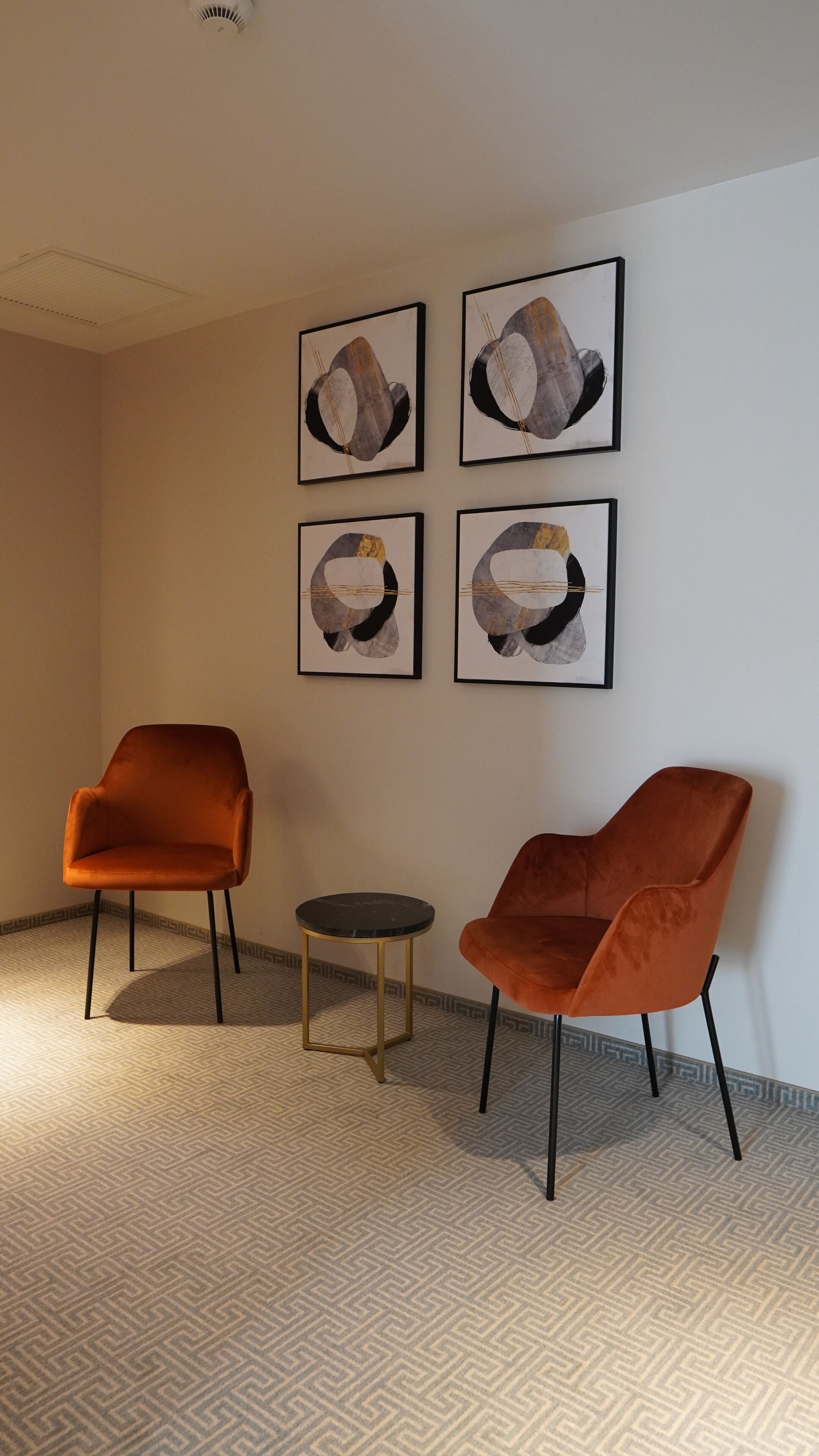 A room with two orange chairs, a small round table, and four abstract paintings on the wall.