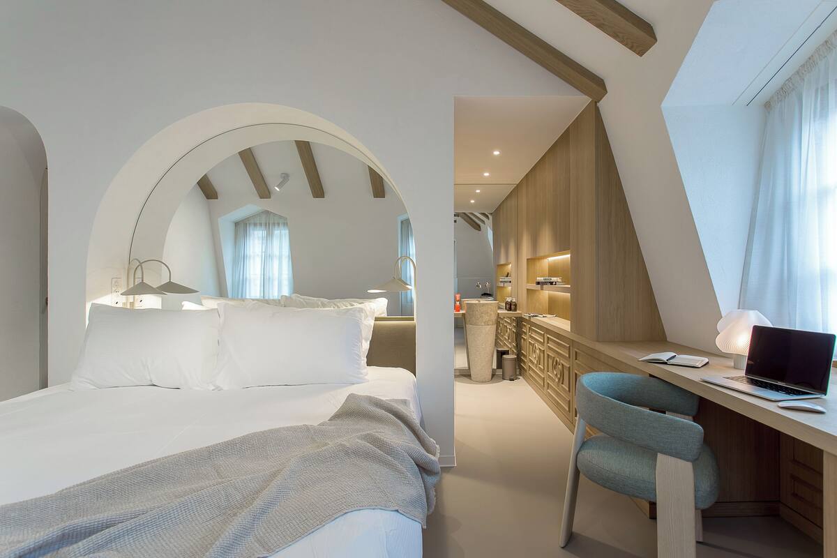 A bedroom with a bed, a desk, and a chair. There is a window with curtains and a view of the outside.