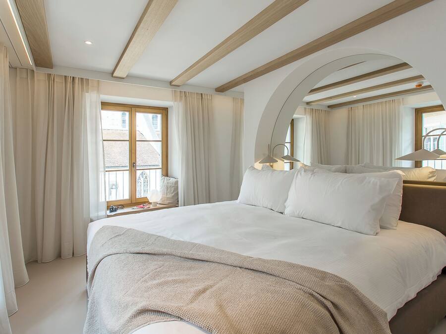 A modern hotel room with a large bed, a window with curtains, a bedside table with a lamp, and a mirror.