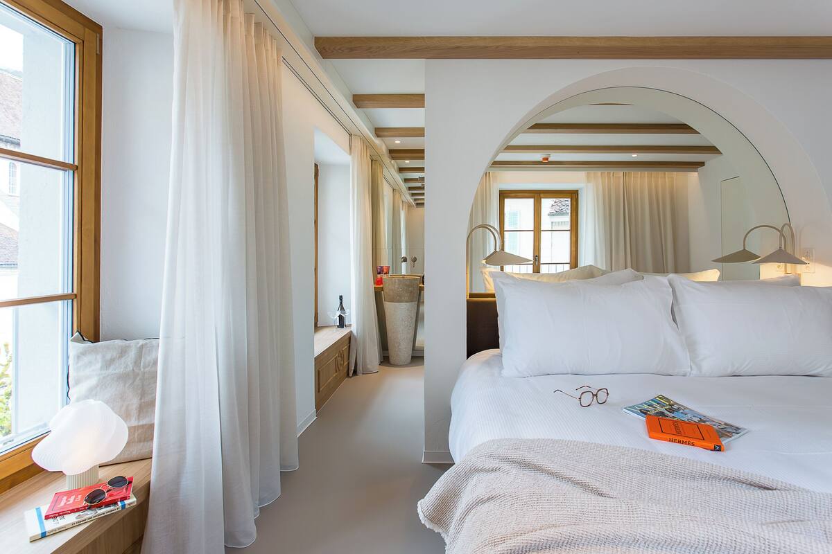 A hotel room with a large bed, a bedside table with a lamp, a window with curtains, and a view of the street.