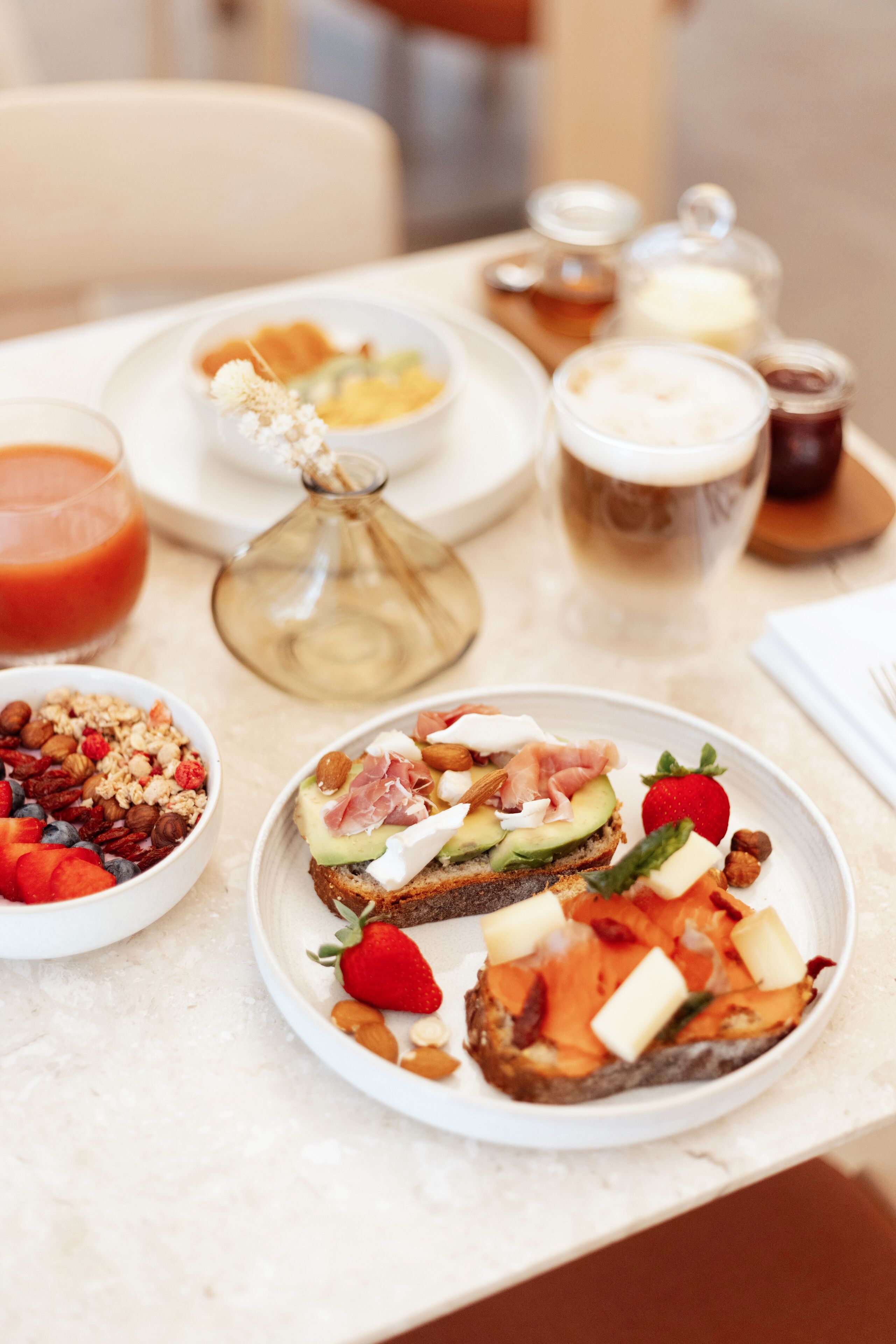 A breakfast setting with various food items on plates, including toast with avocado and smoked salmon, a bowl of granola with fruits, and a glass of orange juice.