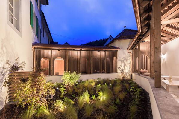 A courtyard with wooden fencing, a covered seating area, and a garden with various plants. - Hotel De la cigogne  (Clos du Doubs)