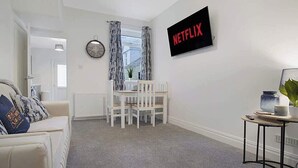 A living room with a white sofa, a dining table with chairs, a clock, a TV with 'NETFLIX' on the screen, and a window with curtains. - Seaside Haven on Newcomen Grove, Redcar Retreat (Redcar and Cleveland)