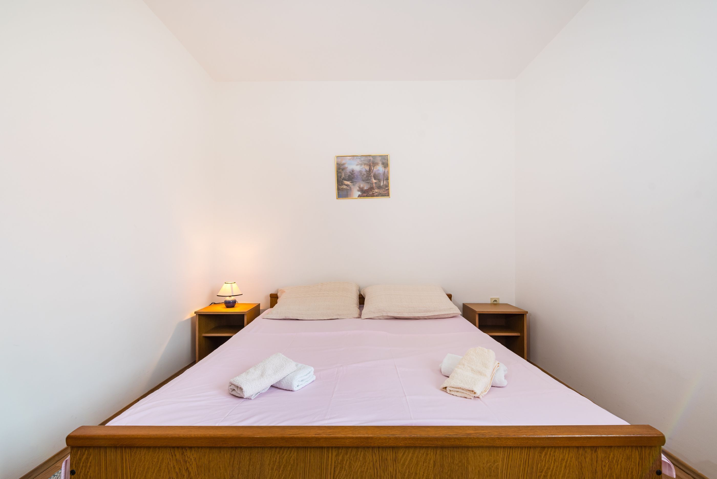 Un lit simple avec un cadre en bois, deux tables de chevet, un tableau accroché au mur et une lampe posée sur l’une des tables.