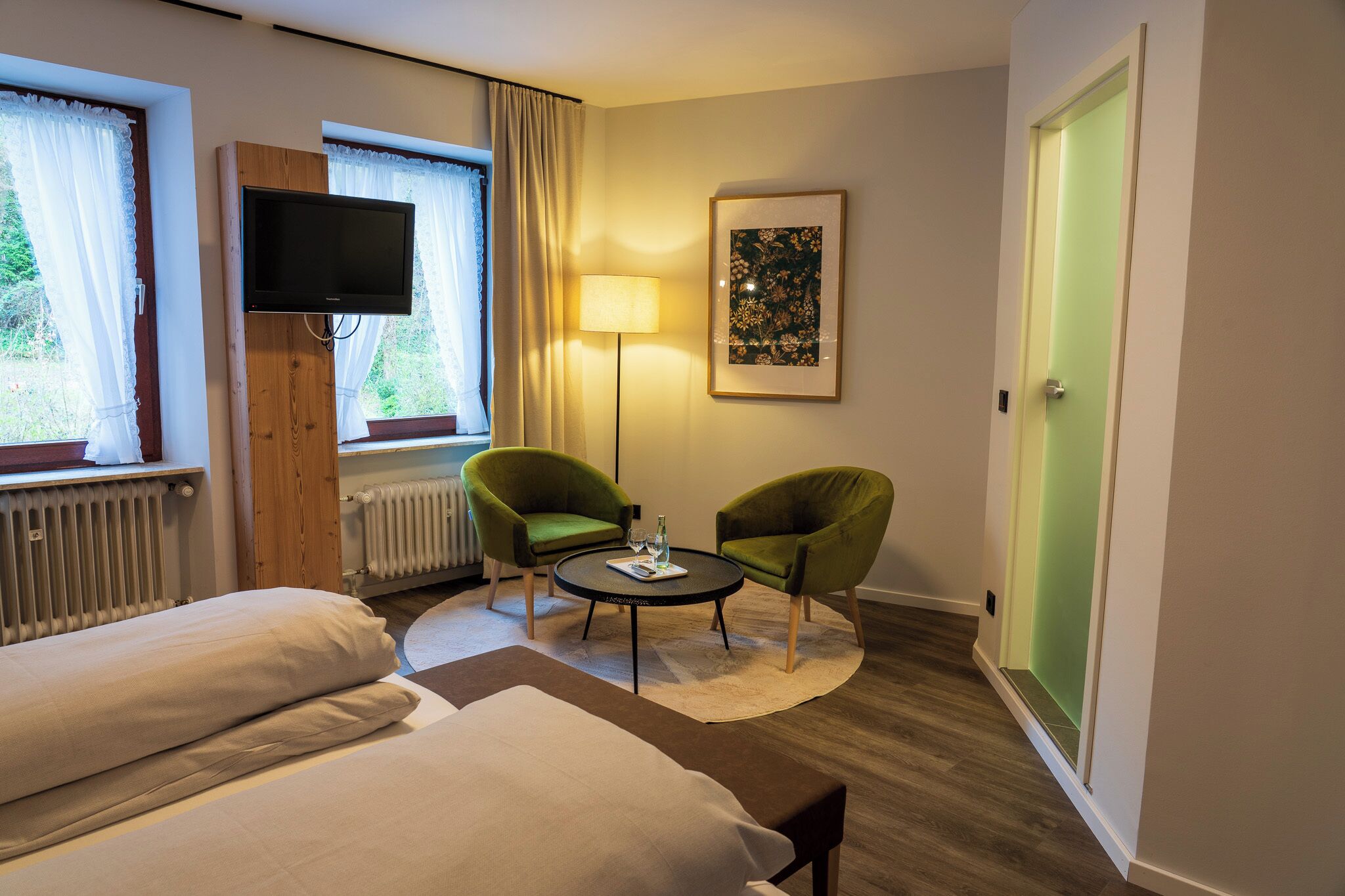 A hotel room with two beds, a flat-screen TV mounted on a wooden panel, a standing lamp, two green armchairs around a small table, and a framed picture on the wall.