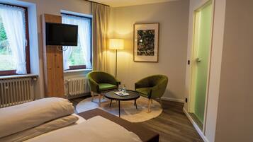 A hotel room with two beds, a flat-screen TV mounted on a wooden panel, a standing lamp, two green armchairs around a small table, and a framed picture on the wall.