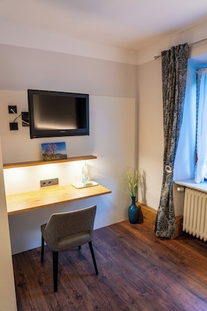 A modern room with a wooden desk, a chair, a TV mounted on the wall, a vase with plants, and a window with patterned curtains. - Hotel Restaurant St. Germanshof (Bobenthal)