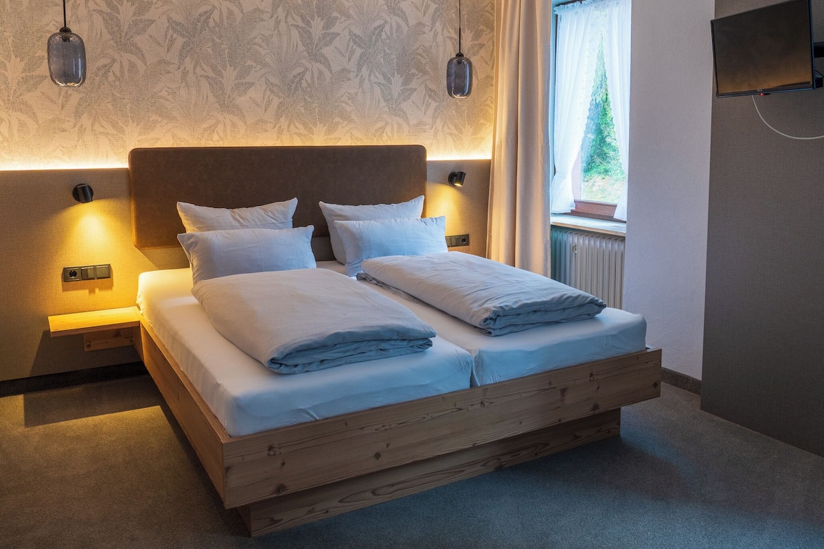 A double bed with a wooden frame, two bedside tables, a wall-mounted TV, and a window with a view of greenery.