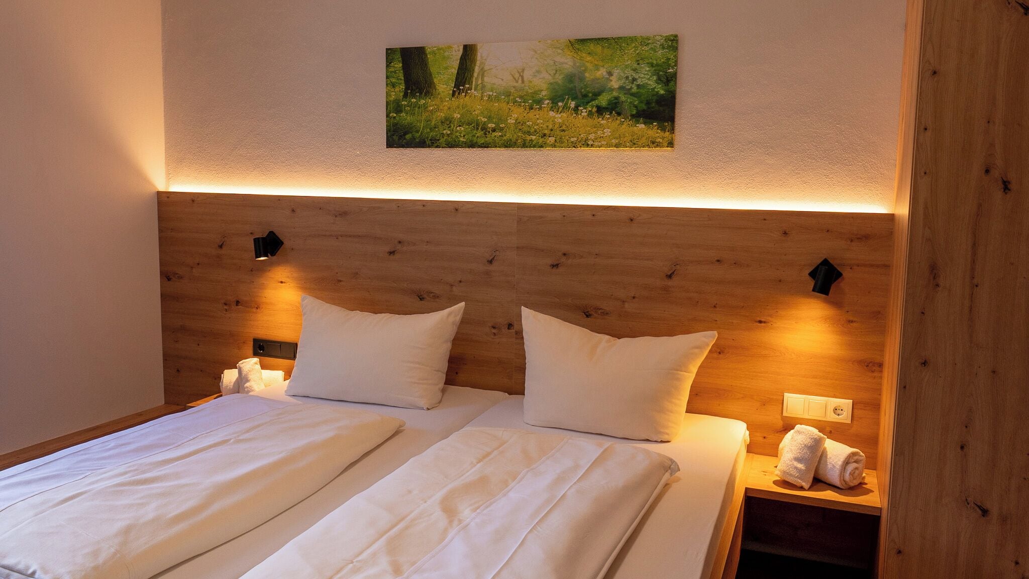 Uma cama de casal com lençóis brancos, cabeceira de madeira, duas luminárias de mesa e um quadro na parede com uma paisagem florestal.