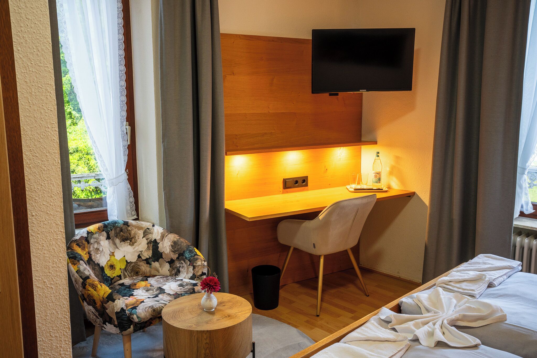 A hotel room with a bed, a desk with a chair, a TV mounted on the wall, and a window with curtains.