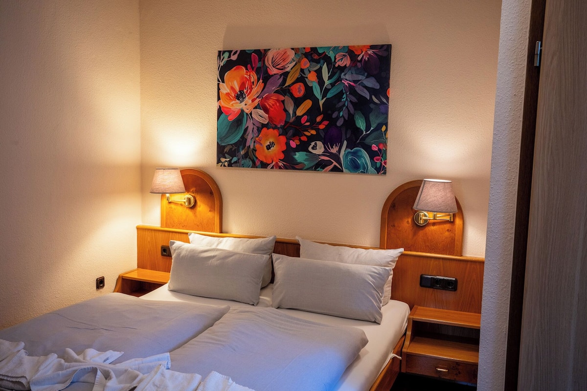 A hotel room with a bed, two bedside lamps, a nightstand, a wooden headboard, and a floral painting on the wall.