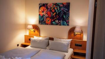 Habitación de hotel con cama, dos lámparas de noche, mesita auxiliar, cabecera de madera y un cuadro floral en la pared.