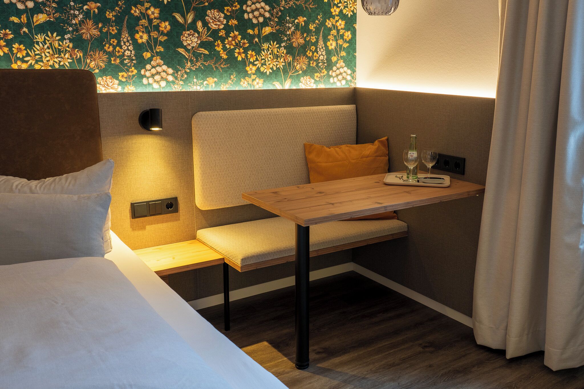 Une pièce confortable avec un lit, un banc agrémenté de coussins, une petite table avec un verre et une bouteille, et un papier peint à motifs floraux.