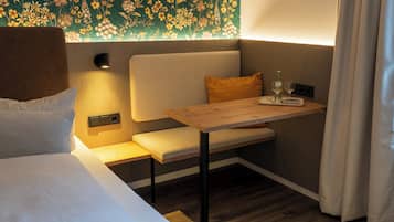 Une pièce confortable avec un lit, un banc agrémenté de coussins, une petite table avec un verre et une bouteille, et un papier peint à motifs floraux.