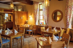 A dining area with wooden tables and chairs, a kitchen in the background, and a round mirror on the wall. - Hotel Restaurant St. Germanshof (Bobenthal)