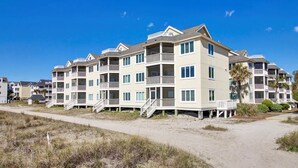 Ein Apartmentkomplex direkt am Strand mit mehreren Balkonen, einem Sandstrand davor und blauem, klarem Himmel.