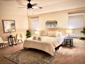 A bedroom with a bed, bedside lamps, a ceiling fan, a mirror, and a window with blinds.