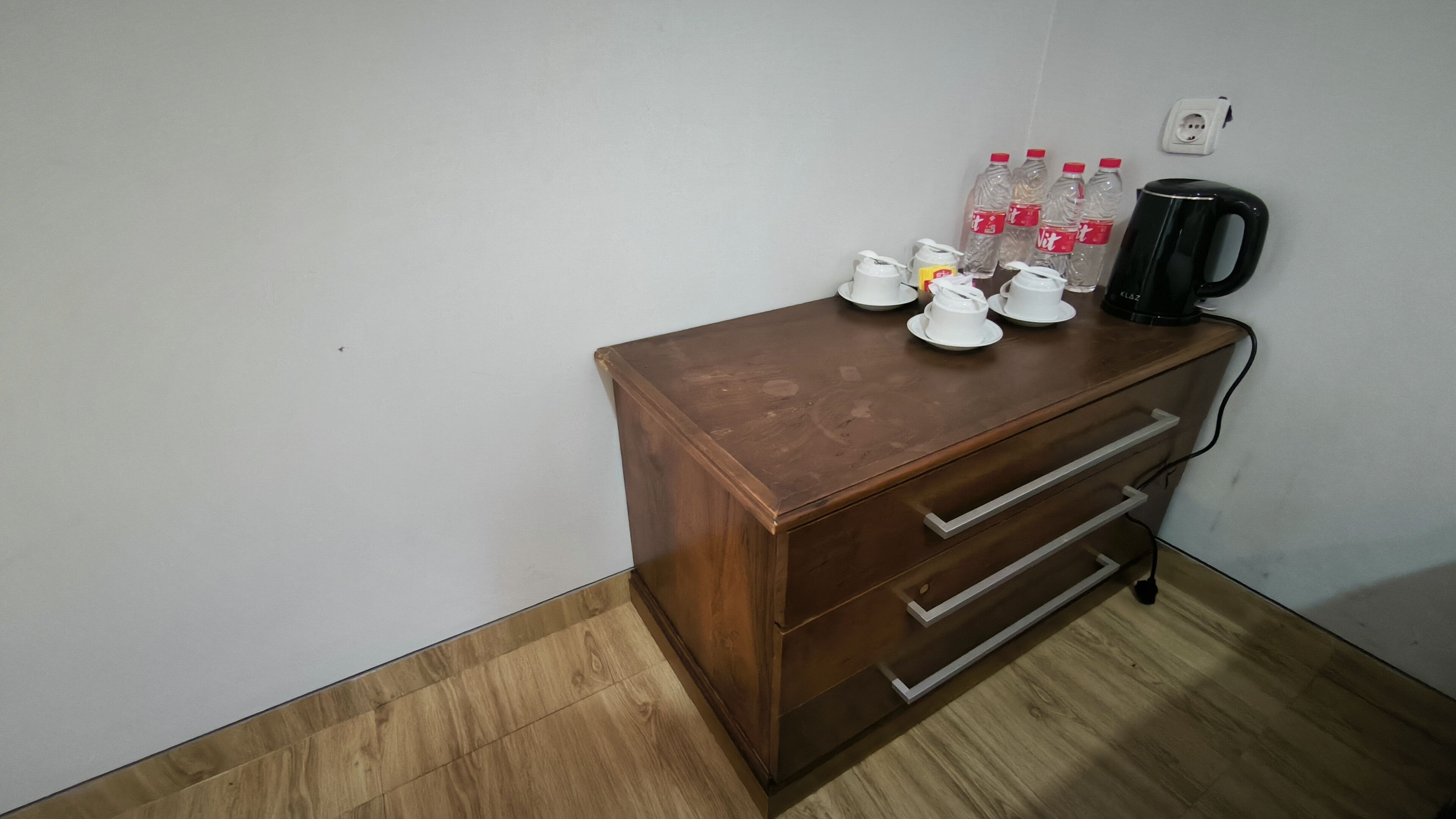 Un pequeño escritorio de madera con cajones, una tetera y varias botellas de agua con vasos.