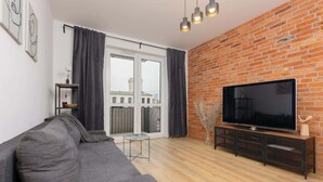 Una sala moderna con un sofá gris, un televisor de pantalla plana sobre un mueble de madera, una pared de ladrillo y un ventanal con cortinas.