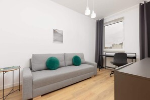 A modern living room with a grey sofa, two green cushions, a small side table, a black office chair, a desk, and a window with curtains.