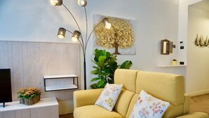 A living room with a yellow sofa, a TV on a wooden cabinet, a large plant, and a wall-mounted shelf with decorative items. - Casa Romano Suites eco apartment AMBRA (Trieste)