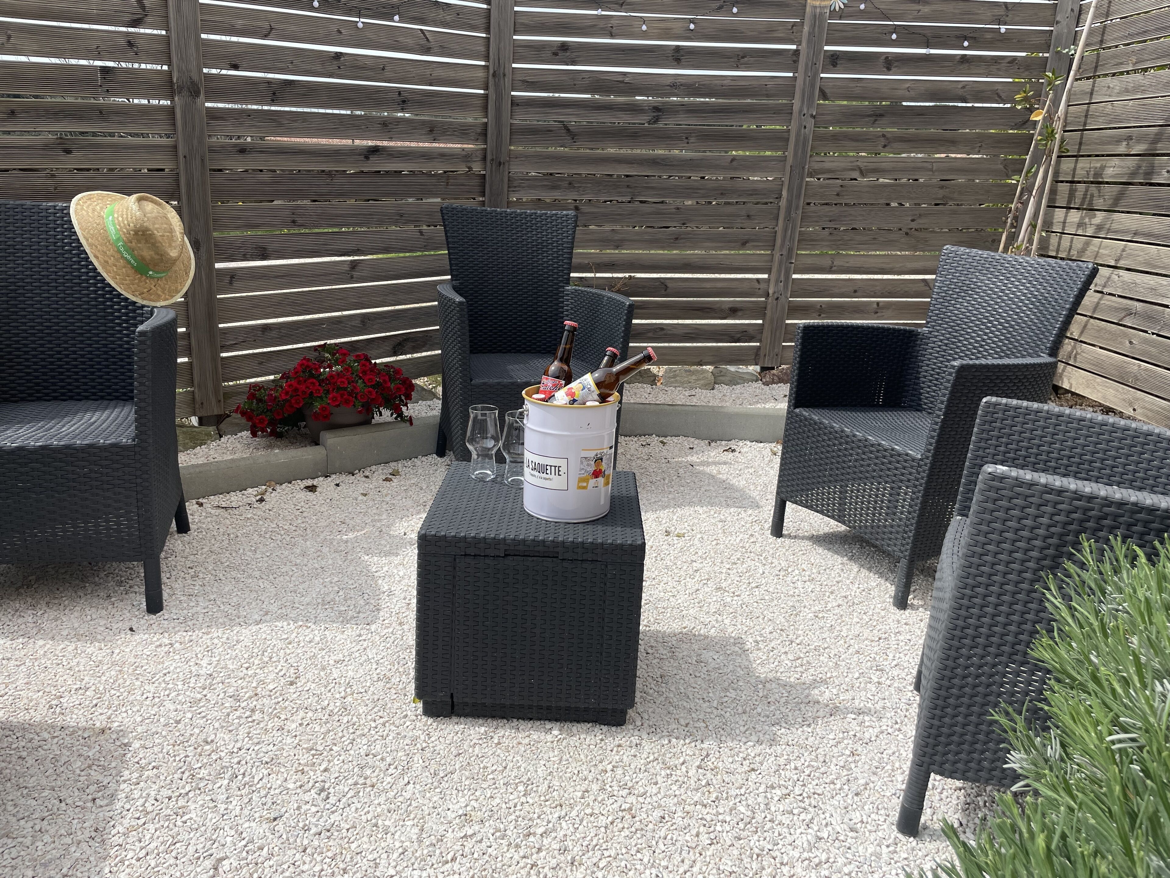 Een terras buiten met rieten meubilair, een witte emmer met de tekst 'LA SAQUETTE' en een biertje.