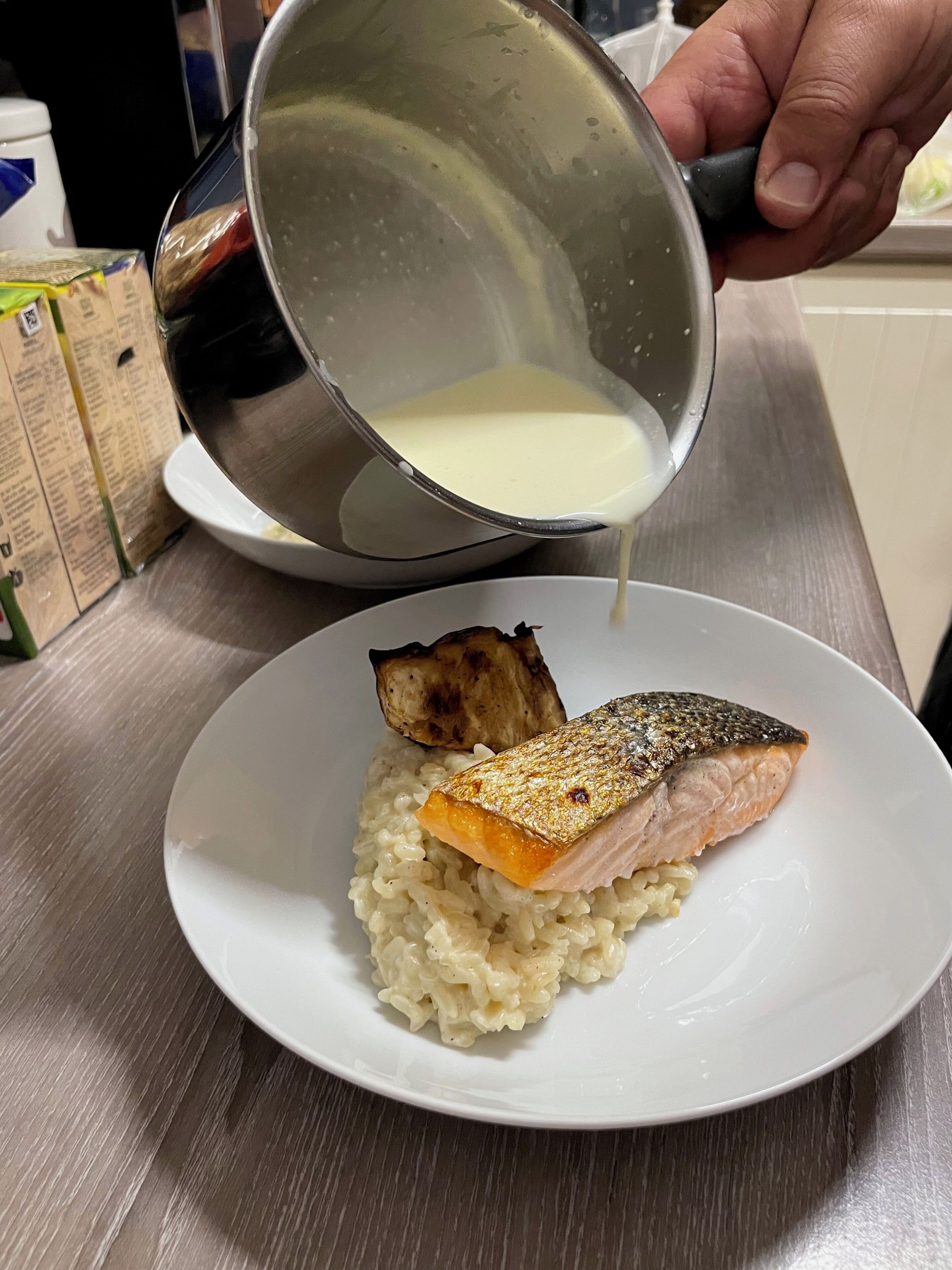 Una persona vertiendo un líquido cremoso en un tazón blanco que contiene arroz y un trozo de pescado.