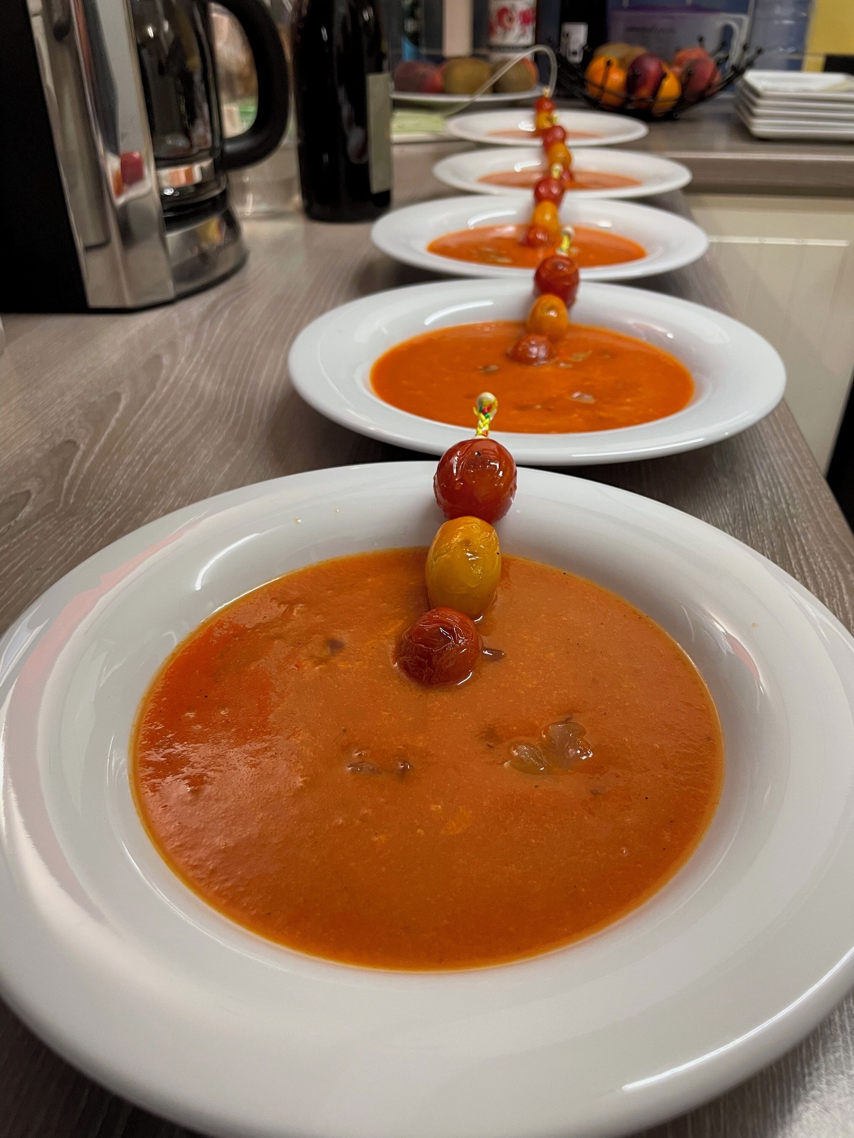 A row of white plates with a red soup garnish, a coffee maker, and a fruit bowl in the background.