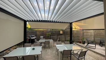 Eine Terrasse im Freien mit überdachter Sitzgruppe, Grill und Blick auf den Garten.