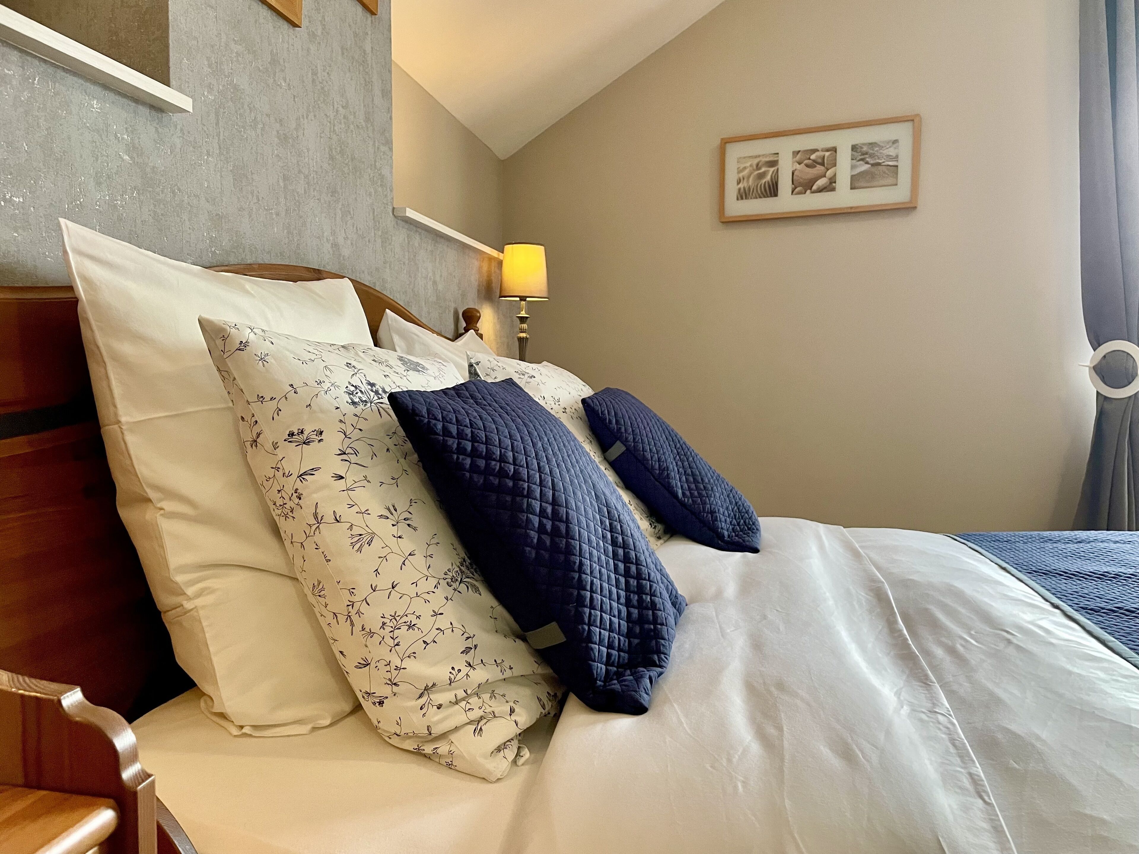 A neatly made bed with floral and quilted pillows, a wooden headboard, a bedside lamp, and a framed picture on the wall.