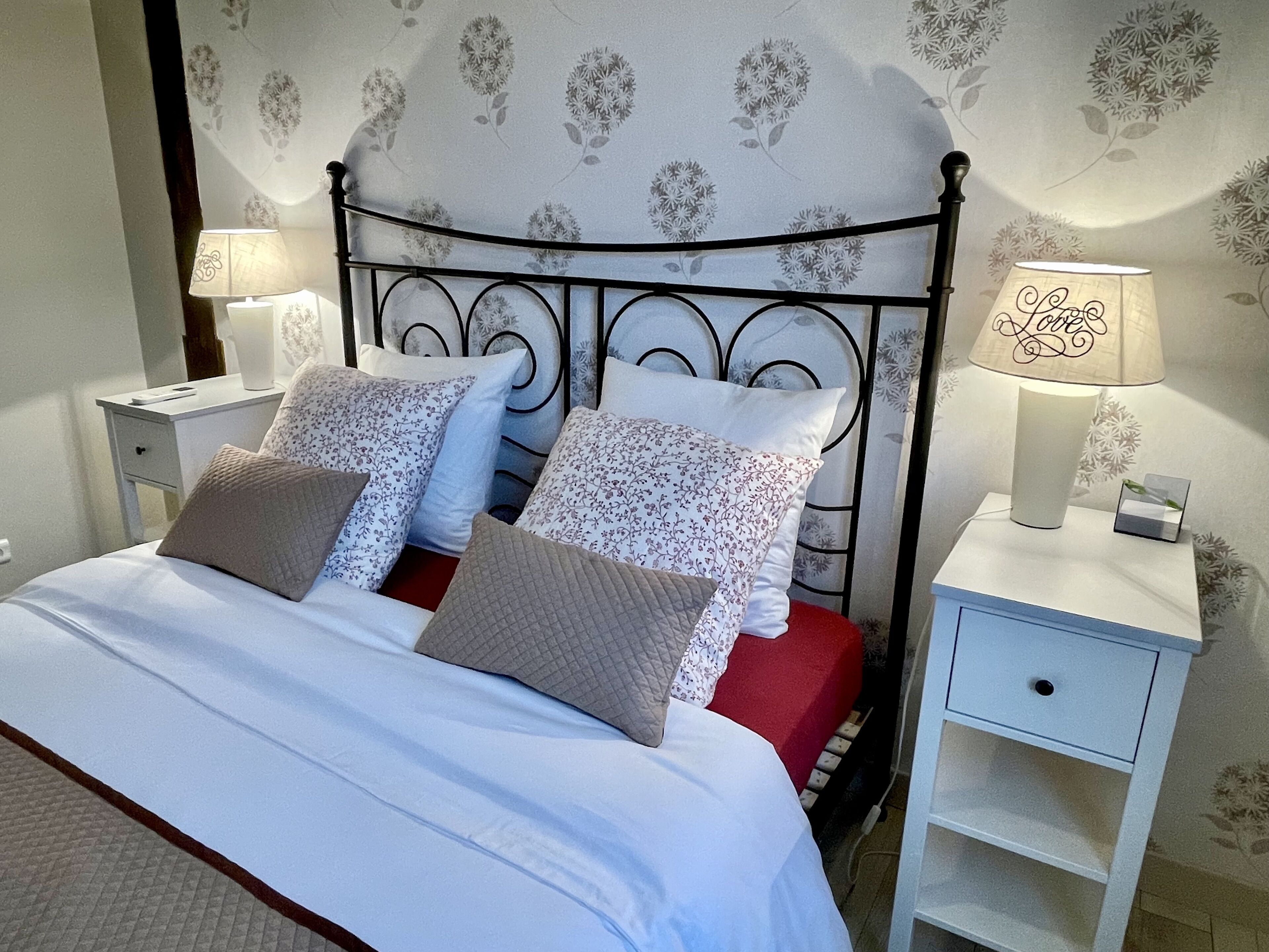 Ein ordentlich gemachtes Bett mit blumigem Kissenmuster, einem Metall-Kopfstück und Nachttischen mit Lampen.