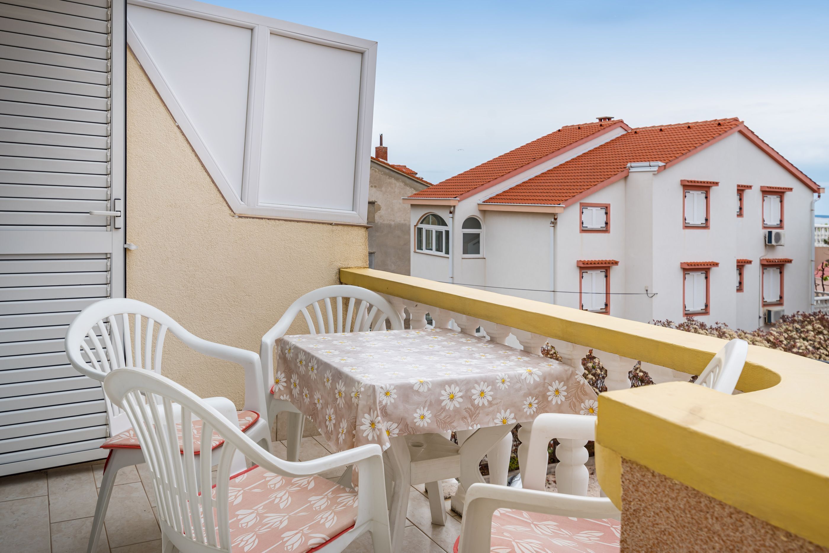 A balcony with a table and chairs, a white door, and a building with red-tiled roofs in the background.