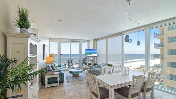 Un balcón amplio con vista a la playa, un comedor con muebles blancos y un televisor montado en la pared.