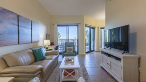 A living room with a beige sofa, a coffee table, a TV on a cabinet, and a balcony with a view. - Phoenix East 008 by Liquid Life (Orange Beach)